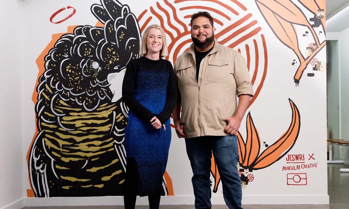 Nicola St John and Emrhan Tjapanangka Sultan stand in front of  a wall painting of a black cockatoo by Jeswri x Mukulri Creative.